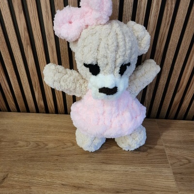 Ursinho de peluche cor creme com saia e laço rosa sobre superfície de madeira