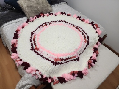 Tapete redondo de lã com pompons em cores branco, rosa e bordô numa cama cinza.
