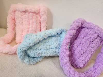 Três malas pequenas de peluche em rosa, azul claro e lilás sobre superfície creme.