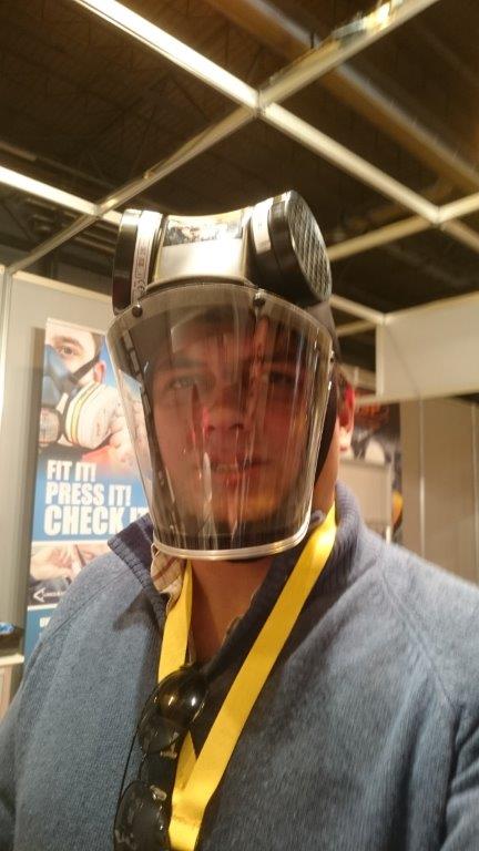 Pessoa com máscara de proteção facial transparente e filtros pretos, fita amarela ao pescoço, em ambiente fechado.