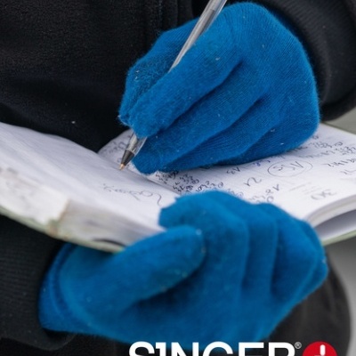 Mãos com luvas azuis escrevendo num caderno, texto SINGER safety no canto inferior direito.