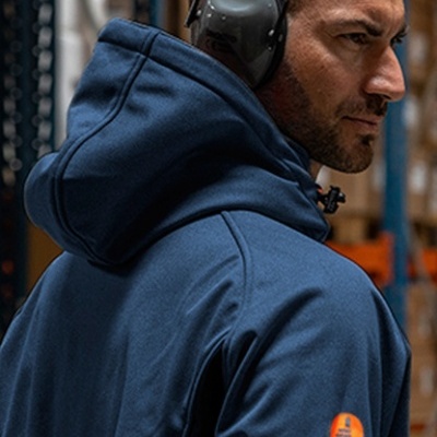 Homem de perfil com casaco azul escuro, protetores auriculares e logótipo laranja na manga, em ambiente industrial