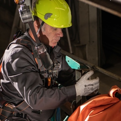 Homem com equipamento de segurança e capacete amarelo em estrutura metálica com sinalização laranja SINGER safety.