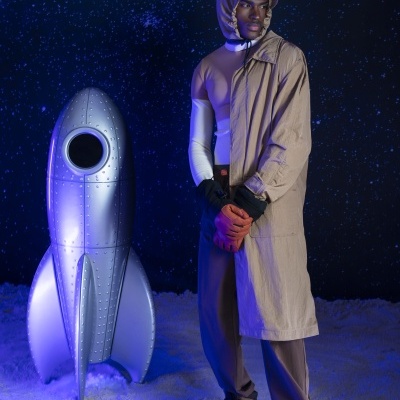 Homem com casaco bege e botas ao lado de foguete prateado no espaço azul