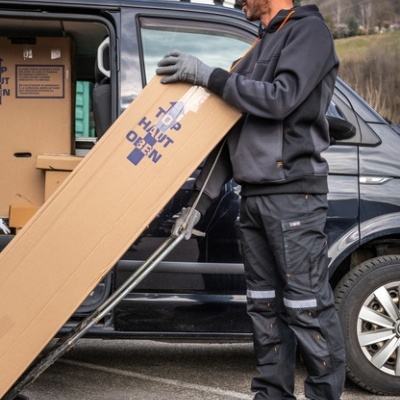 Homem a transportar caixa de cartão longa junto a carrinha preta com logotipo SINGER safety