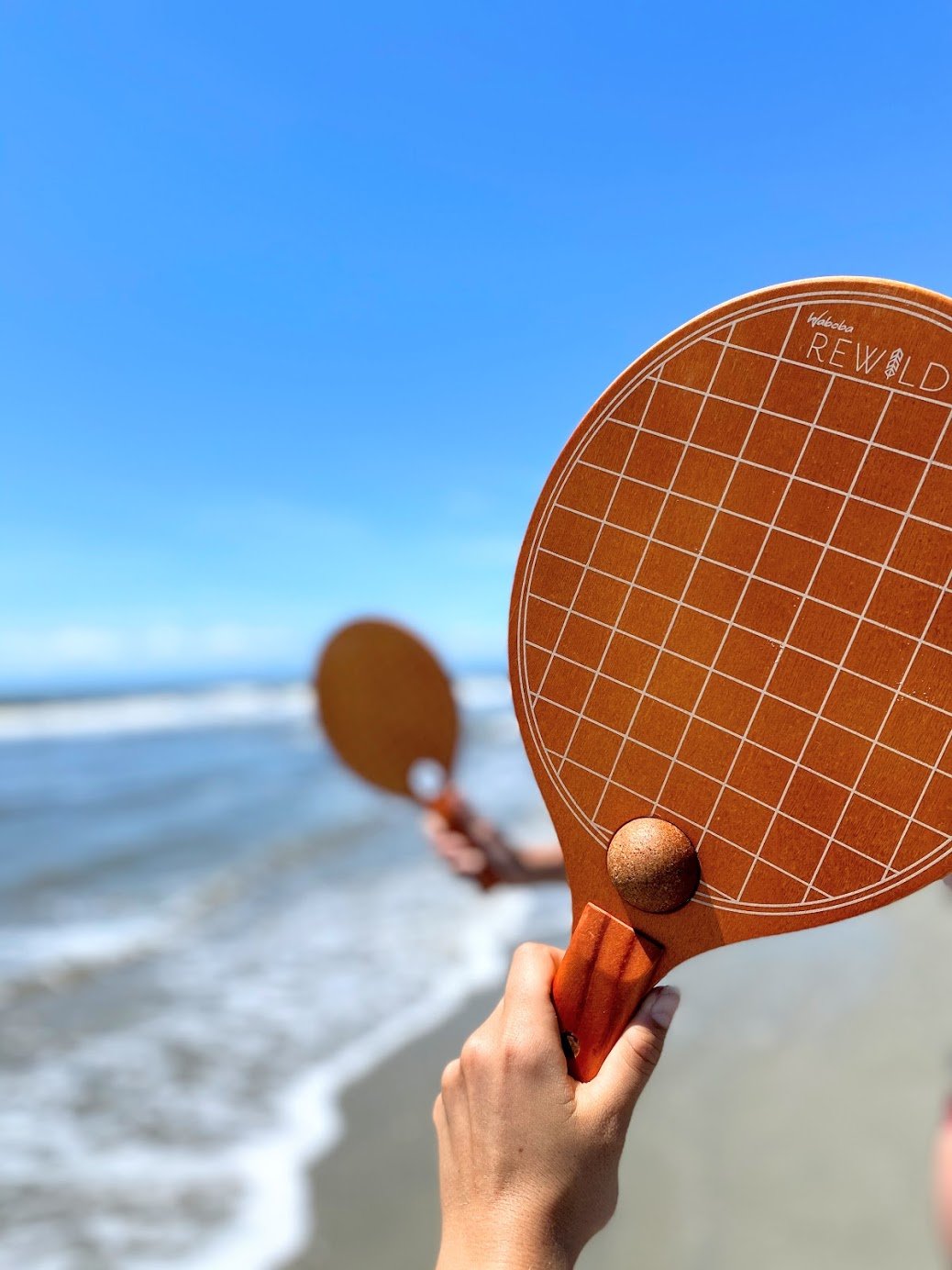 Raquetes Paddle Rewild Raquete de madeira para jogo de praia com padrão de grelha branca e bola castanha na mão, com praia e mar ao fundo