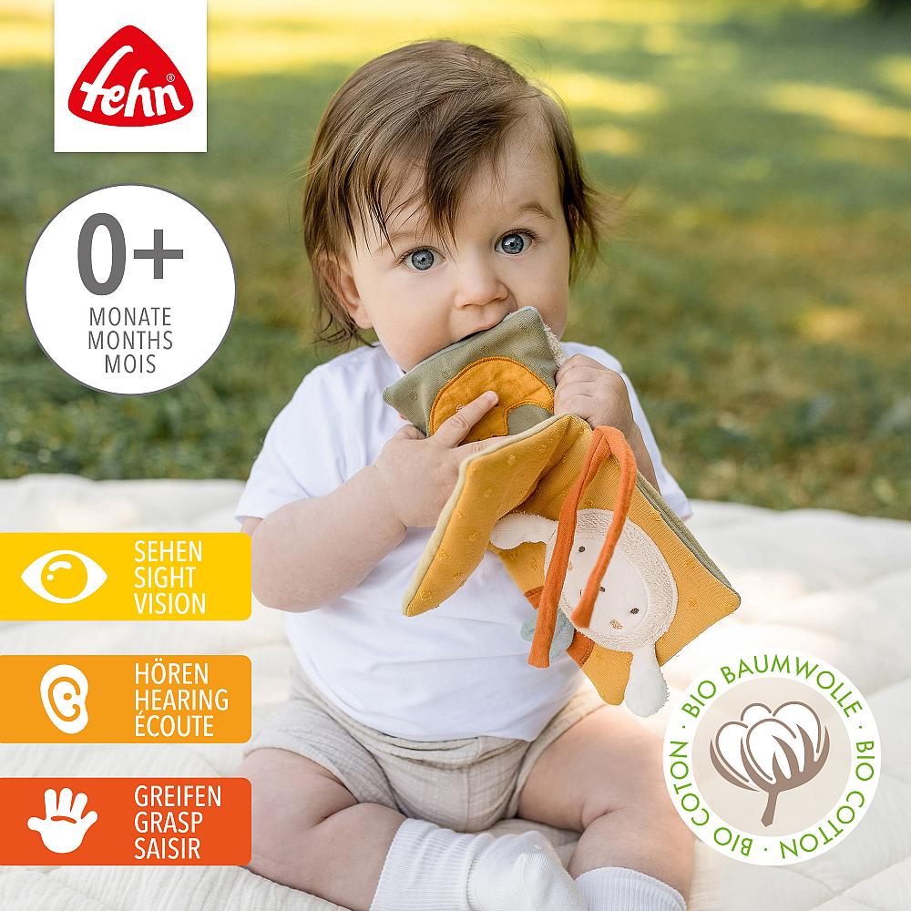Livro Suave ovelha fehnNATUR Bebé a brincar com brinquedo macio em tecido colorido sentada ao ar livre