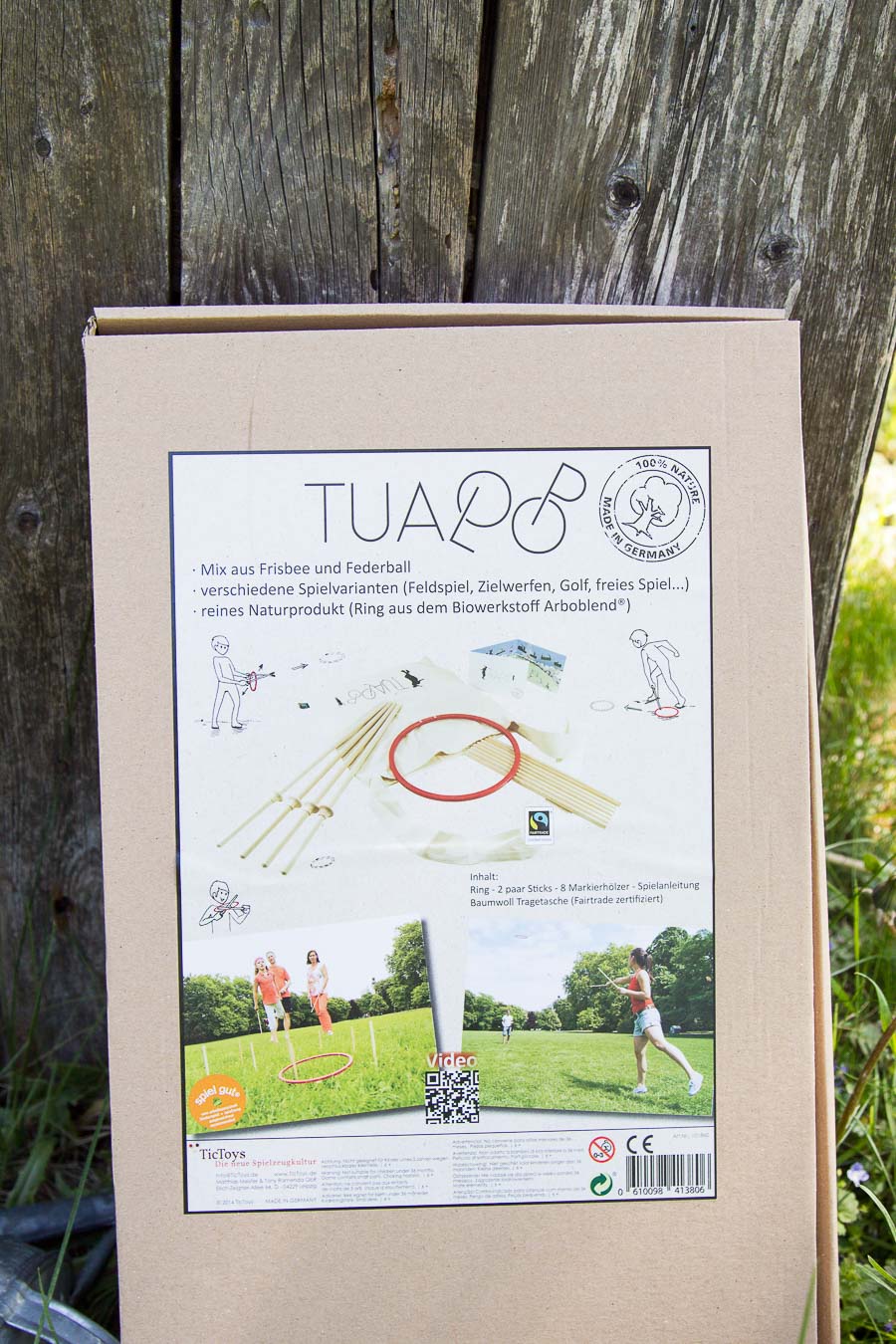 Jogo Tualoop Caixa de produto de jogo ao ar livre com imagens e texto em alemão, encostada a uma cerca de madeira