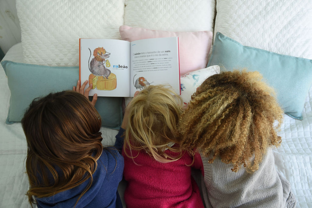 Três crianças de cabeça baixa a ler um livro infantil numa cama com almofadas coloridas