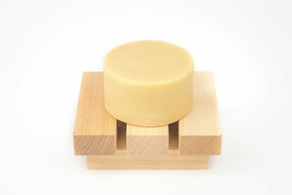 AMACIADOR CONDICIONADOR SÓLIDO Sabonete redondo bege num suporte de madeira clara com três ripas em fundo branco