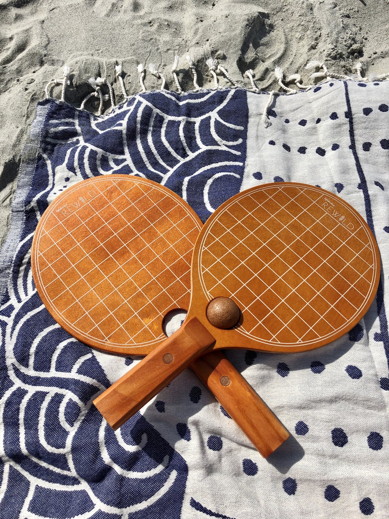 Raquetes Paddle Rewild Duas raquetes de madeira sobre toalha de praia azul e branca na areia