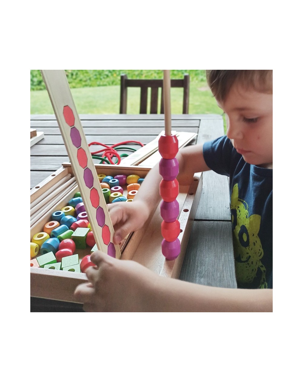 Formas para Encaixe - 92 Peças Criança a jogar com contas coloridas de madeira para empilhar e cartão com padrões coloridos numa caixa de madeira.