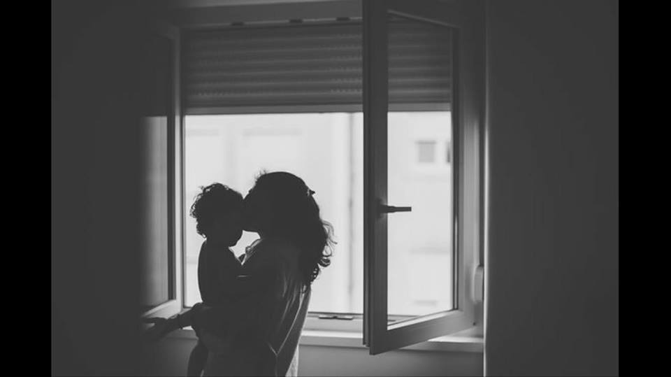 Silhueta de mulher a segurar criança junto a janela aberta com persiana
