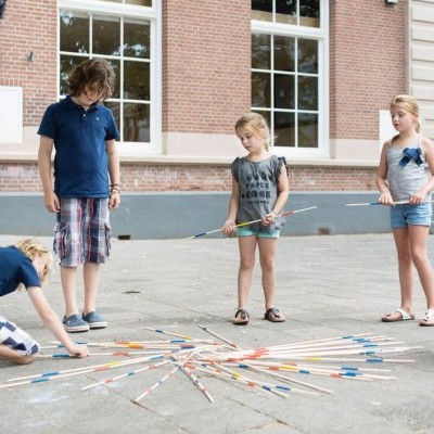 Crianças a jogar Mikado gigante ao ar livre, bastões coloridos no chão de cimento em frente a prédio de tijolos.