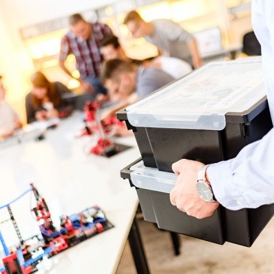 Caixa preta com tampa translúcida sendo transportada numa sala de aula com pessoas e robôs na mesa