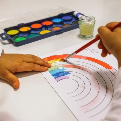 Criança a pintar um arco-íris com aguarelas coloridas num papel branco.