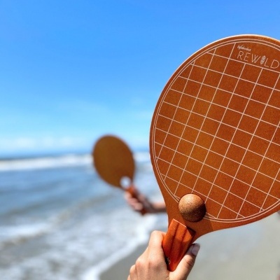 Raquete de madeira para jogo de praia com padrão de grelha branca e bola castanha na mão, com praia e mar ao fundo