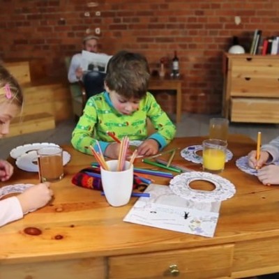 Crianças a desenhar numa mesa de madeira com materiais de arte, homem sentado num sofá ao fundo