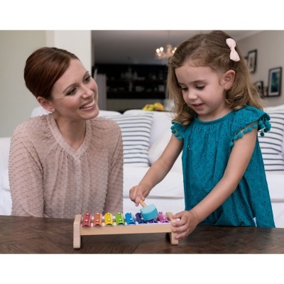 Menina a tocar xilofone colorido de madeira com mãe a sorrir sentado num sofá em casa.