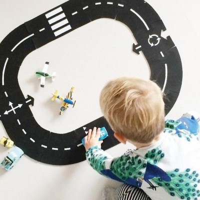 Criança a brincar com pista de corrida preta e carros e aviões de brinquedo coloridos sobre superfície branca