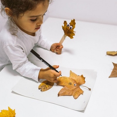 Criança a decorar folhas secas com olhos móveis e a desenhar num caderno branco sentado numa mesa branca