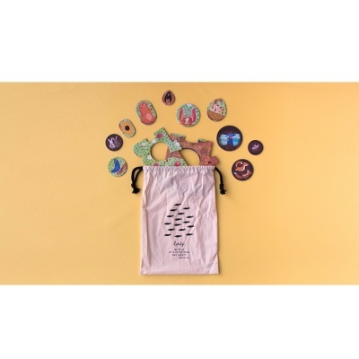 Saco tecido bege com ilustração de peixes e vários marcadores coloridos sobre fundo amarelo.