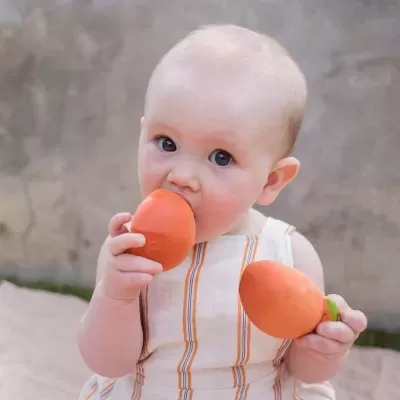 Bebé segurando brinquedos laranja em forma de cenoura, roupa branca às riscas laranjas, fundo cinzento desfocado