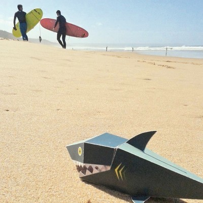 Boneco dobrável de tubarão na areia da praia com surfistas ao fundo