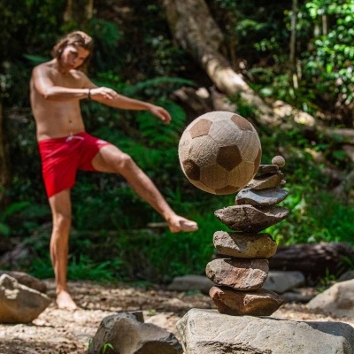 Bola de futebol castanha e bege em padrão hexagonal perto de pedras empilhadas numa floresta com homem a chutar.