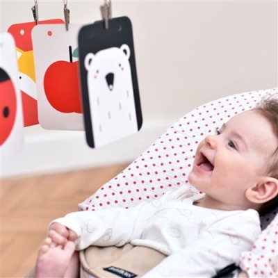 Bebé sorrindo sentado em cadeira com almofada branca com pontos vermelhos, olhando cartões coloridos pendurados