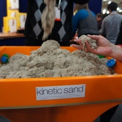 Caixa laranja com areia cinética beje e etiqueta 'kinetic sand', mão segurando areia em ambiente interno