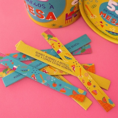 Pulseiras de tecido coloridas e latas com texto sobre desafios à mesa, fundo rosa.