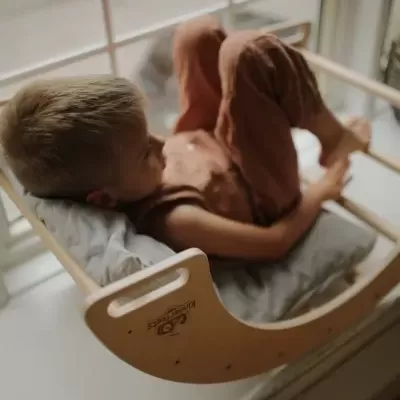 Cadeira de baloiço em madeira clara com almofada cinza e uma criança de roupa castanha a descansar