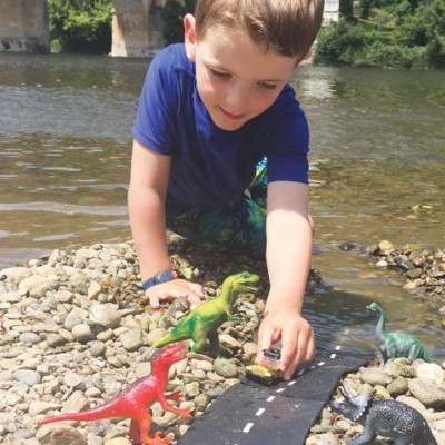 Criança a brincar com dinossauros de plástico numa margem de rio com seixos e estrada de brincar.