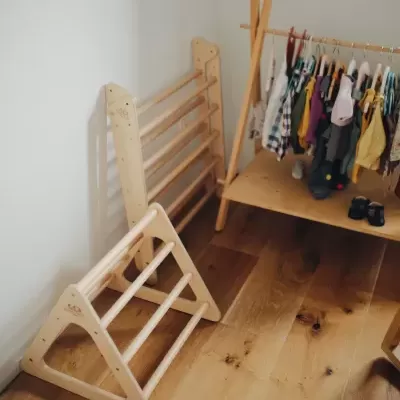 Estrutura triangular de madeira clara com barras, suporte vertical, cabideiro com roupas e sapatos no chão de madeira