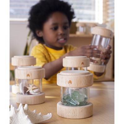 Frascos de vidro com tampa e base de madeira clara contendo conchas e pedras sobre mesa com criança ao fundo