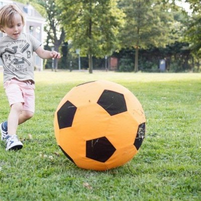 Bola insuflável de futebol laranja e preta em relvado verde com criança a jogar.