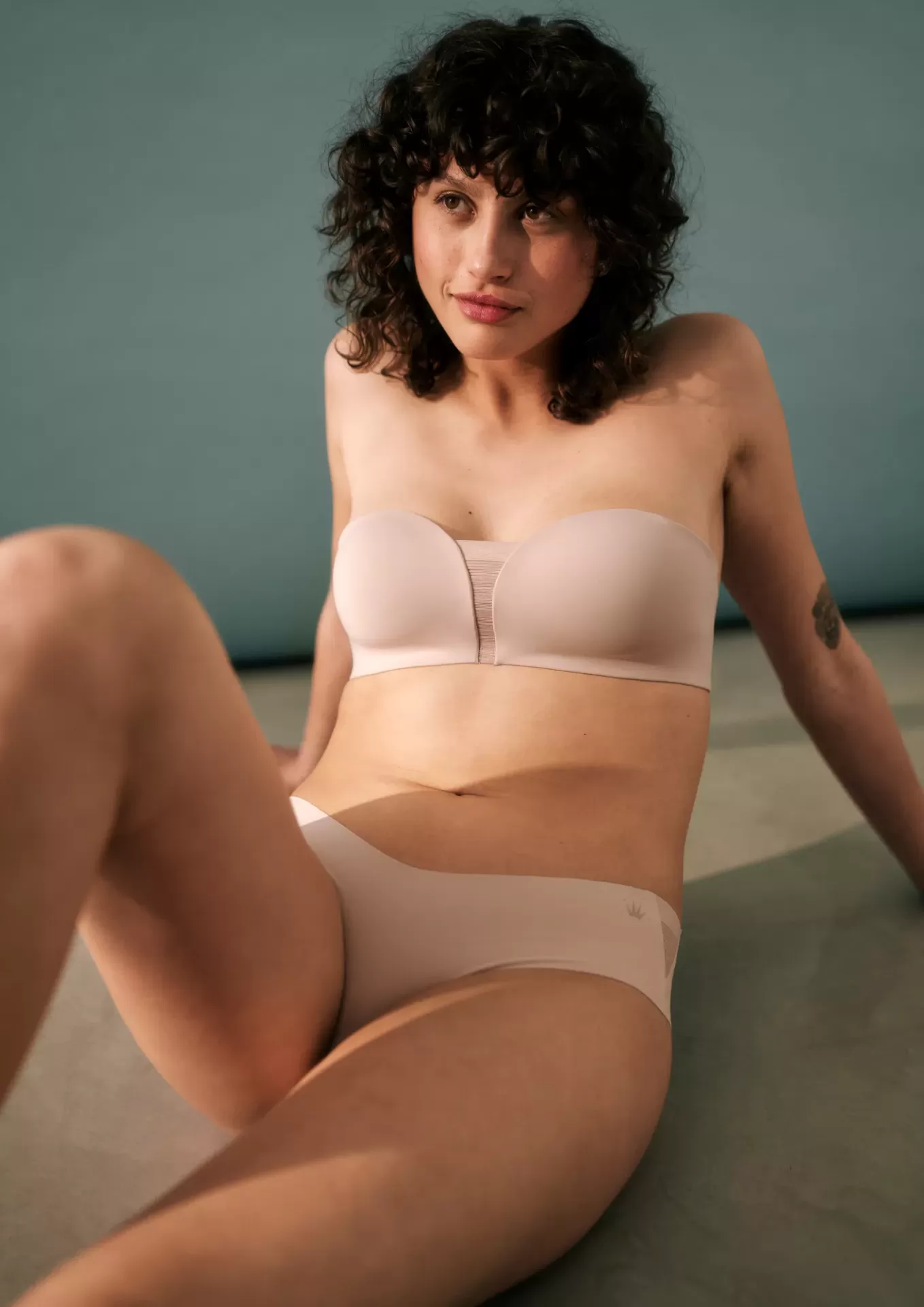 Mulher com conjunto de roupa interior cor pele, sutiã tomara que caia e cueca, sentada no chão com parede cinzenta ao fundo