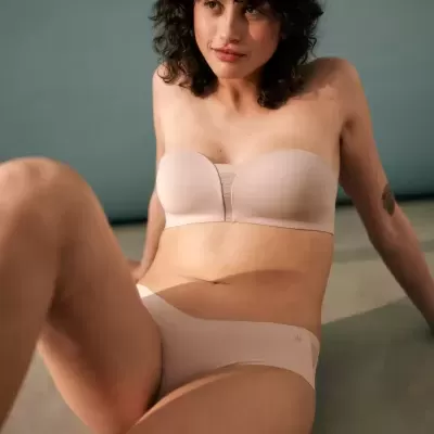 Mulher com conjunto de roupa interior cor pele, sutiã tomara que caia e cueca, sentada no chão com parede cinzenta ao fundo