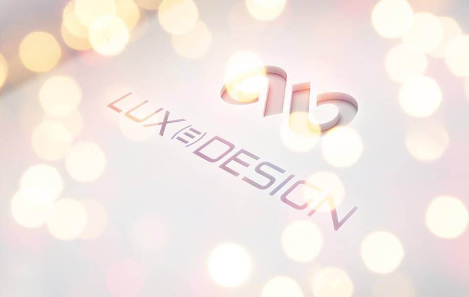 Logótipo e texto 'LUX DESIGN' em tons dourados e rosa sobre superfície branca com luzes desfocadas