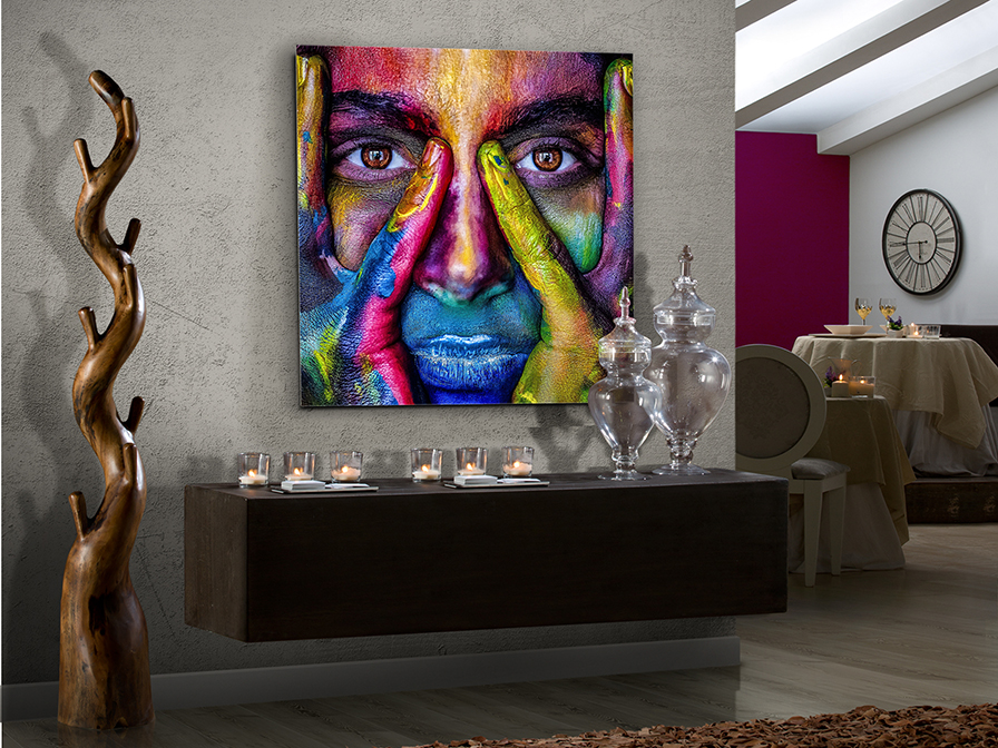 COLOURS Quadro colorido de rosto humano pintado em várias cores vibrantes numa parede cinzenta acima de mesa preta com velas e objetos de vidro num interior moderno.