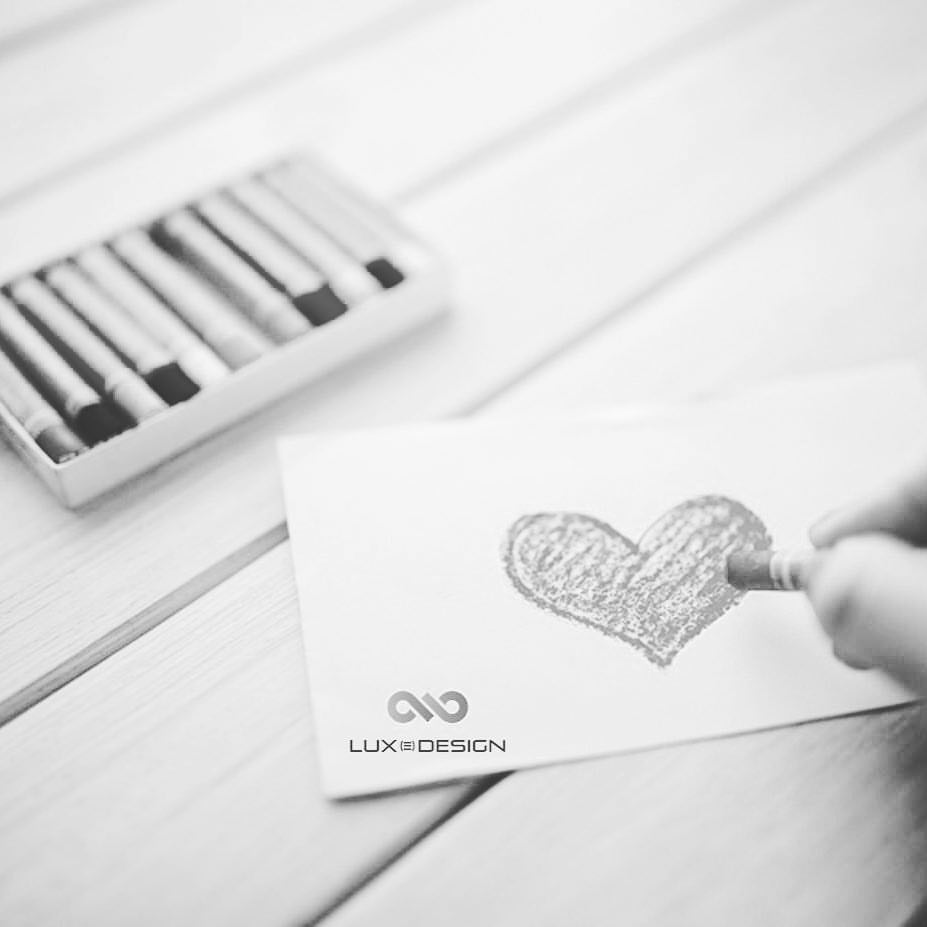 Mão a desenhar coração com lápis de cor, caixa de lápis ao lado, papel e madeira clara