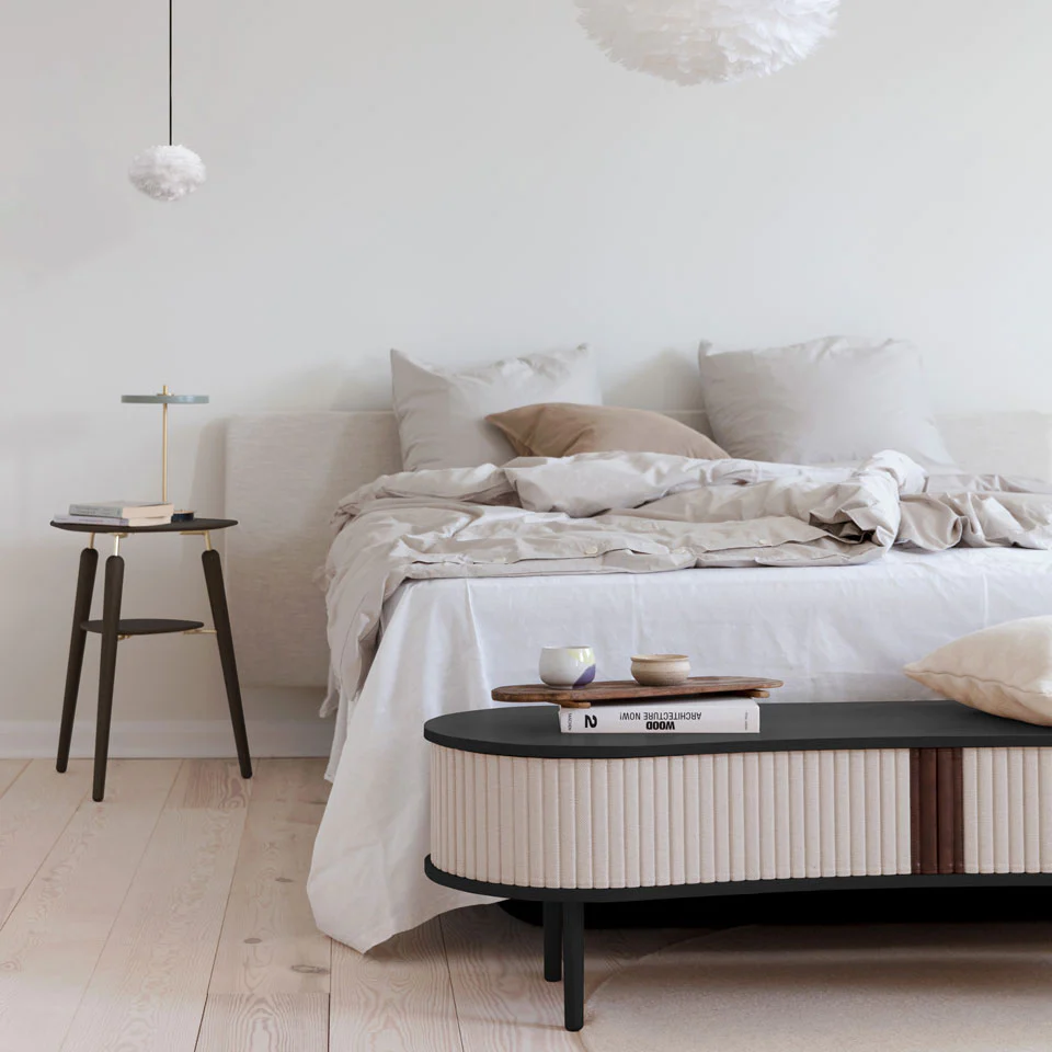 Quarto minimalista com cama branca, almofadas bege, mesa de cabeceira preta e mesa de centro oval preta e bege.