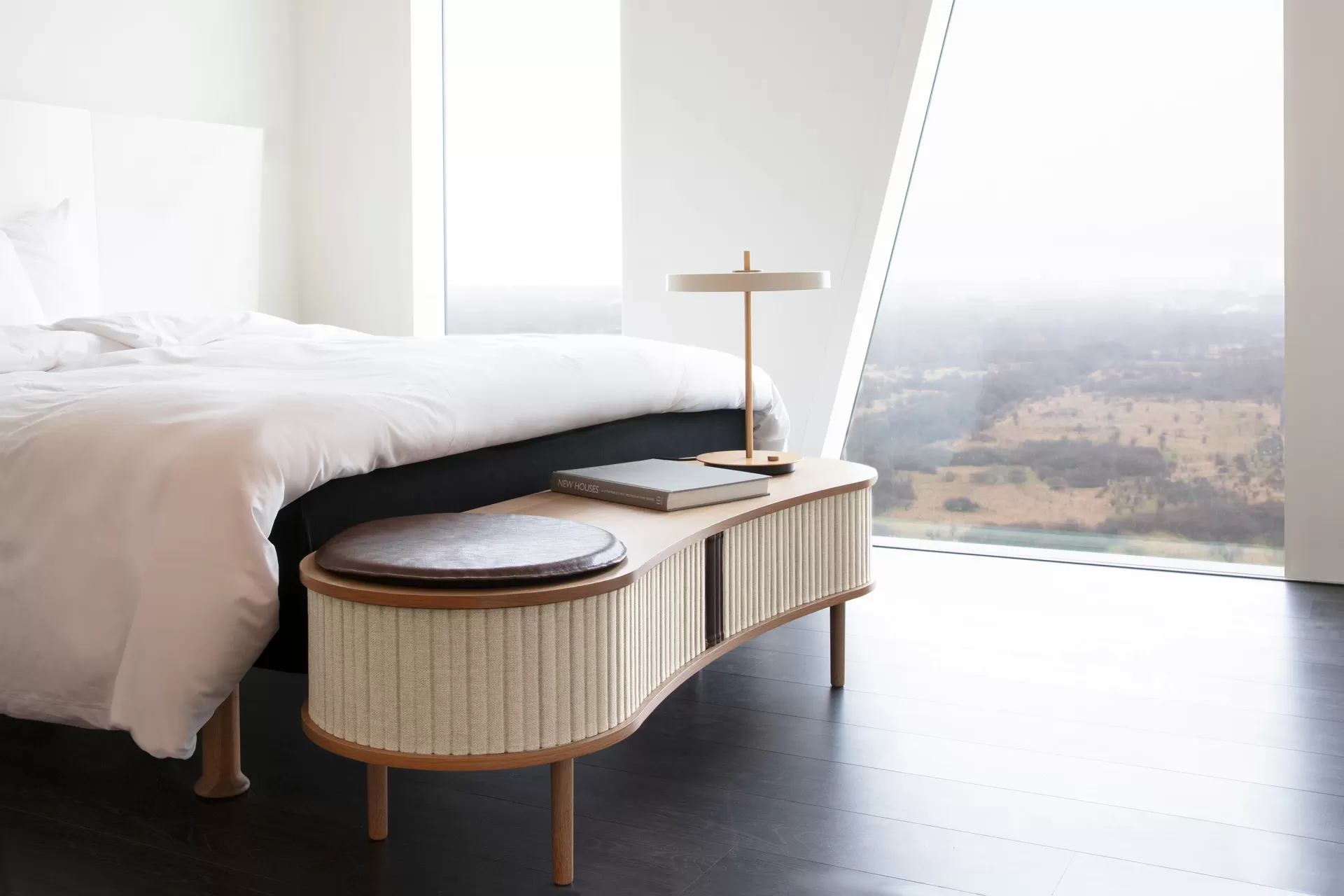 Quarto moderno com cama branca e mesa de cabeceira de madeira junto a janela ampla.