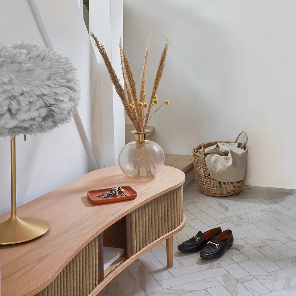 Área de entrada com consola de madeira clara e decoração com vaso, candeeiro, sapatos e cesto de palha