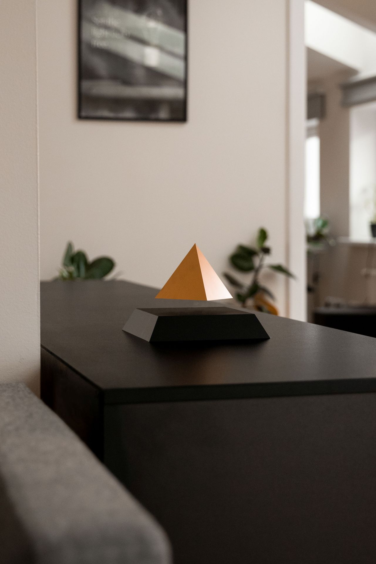 FLYTE PY Escultura de pirâmide dourada com base preta sobre superfície preta em interior com parede clara