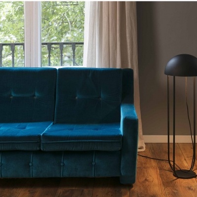 Sofá de veludo azul com botões, candeeiro de pé preto, janela com cortinas brancas