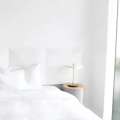 Quarto com cama branca, mesa-de-cabeceira cinzenta e candeeiro com base preta e haste dourada