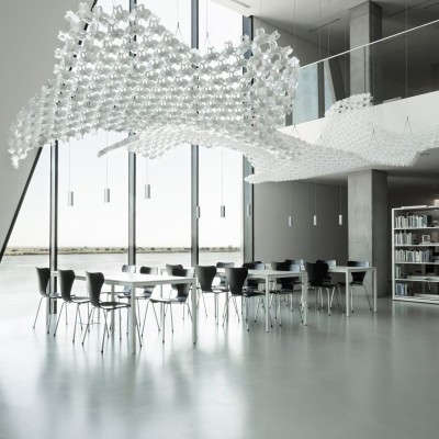 Sala moderna com mesas, cadeiras pretas, luminárias brancas suspensas e estantes de livros