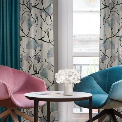 Sala com cadeiras rosa e azul, mesa redonda com vaso branco e cortinas florais.